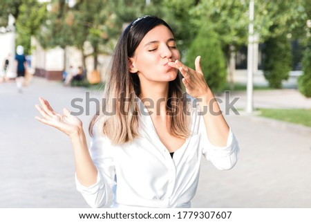 portrait of beautiful woman in white shirt and hair bezel with mouth full of food licking her fingers outdoor in city park and enjoying junk but tasty fast food while walking