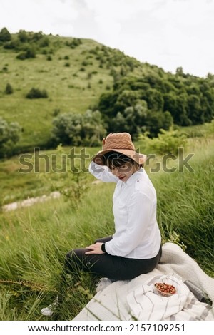 The portrait of beautiful woman in white shirt and brown hat sitting on plaid, green grass, wild ripe red strawberries in summer mountain. Nice nature, cozy, healthy fruit, travel, village.