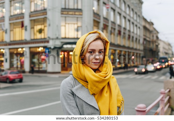 ショーウィンドウの背景に金髪の美しい女性のポートレート 黄色いスカーフとグレーコートを着て 通りのスタイル 幸せな女性の接写 の写真素材 今すぐ編集
