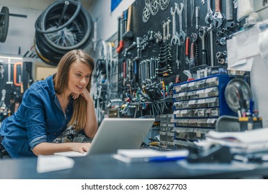 Portrait of beautiful working woman in the bicycle store. Young female mechanic with laptop. Female Bicycle Mechanic. Craftswoman working using a laptop in the garage - Powered by Shutterstock