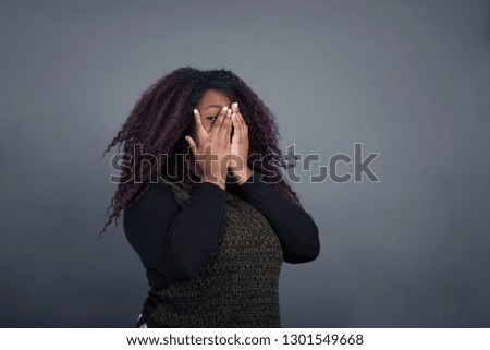 Portrait of beautiful young African American woman covering her face with her hands and peering out with one eye between her fingers. Scared from something or someone. Standing against gray wall.
