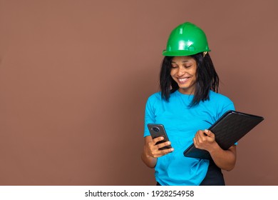 Portrait Of A Beautiful Young African Female Construction Worker Wearing A Heard Hat, Using Her Mobile Phone, Carrying A Document, Standing Against A Plain Background