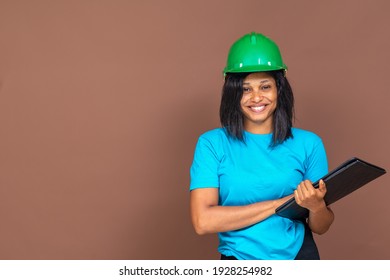 Portrait Of A Beautiful Young African Female Construction Worker Wearing A Heard Hat, Carrying A Document, Standing Against A Plain Background