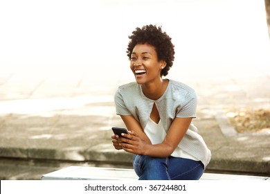 Portrait of beautiful young african woman smiling while sitting outside on a bench holding mobile phone
