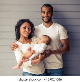Portrait of beautiful young Afro American parents looking at camera and smiling, standing against gray wall. Little baby is sleeping in mom's arms