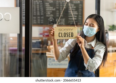 Portrait Of A Beautiful Young Asian Barista Wearing An Apron. Holding A Sign With The Word Closed At The Glass Door Working At A Coffee Shop.