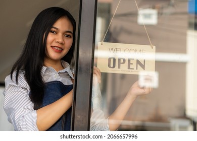 Portrait Of A Beautiful Young Asian Barista Wearing An Apron. Holding An Open Sign At The Glass Door Of The Cafe.