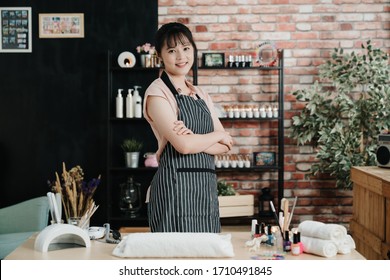 Portrait Of Beautiful Young Asian Chinese Woman Manicurist Standing In Front Of Nail Salon And Smiling Face Camera. Confident Beautician Girl Wear Striped Apron Crossed Arms In Beauty Workshop Indoor