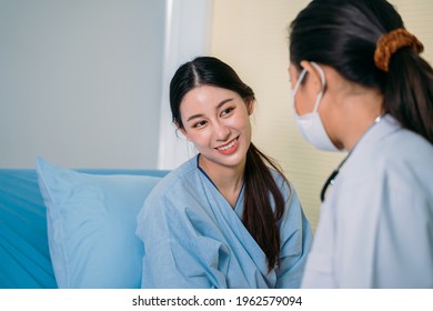 Portrait of beautiful young Asian female patient in scrubs talking to doctor wearing face mask for safety while sitting on hospital bed during consultation - Powered by Shutterstock