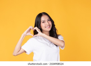 Portrait of beautiful young Asian woman model long hair in casual white shirt smile, happy and enjoying in studio isolated on yellow background. The model raises her hand as an heart symbol. - Powered by Shutterstock