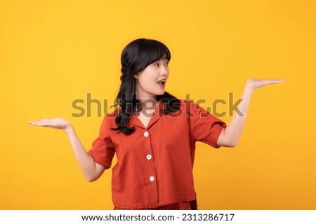Portrait beautiful young asian woman happy smile dressed in orange clothes showing hand gesture, arm compare two variants demonstrate empty space isolated on yellow studio background.