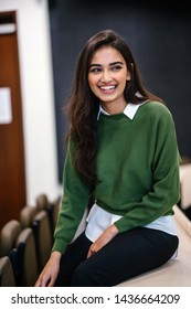 Portrait Of A Beautiful, Young And Attractive Indian Asian Business Woman Smiling As She Sits In A Seminar Room During The Day. She Is Wearing A Preppy Green Sweater Over A White Shirt And Jeans. 