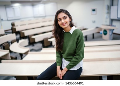 Portrait Of A Beautiful, Young And Attractive Indian Asian Business Woman Smiling As She Sits In A Seminar Room During The Day. She Is Wearing A Preppy Green Sweater Over A White Shirt And Jeans. 