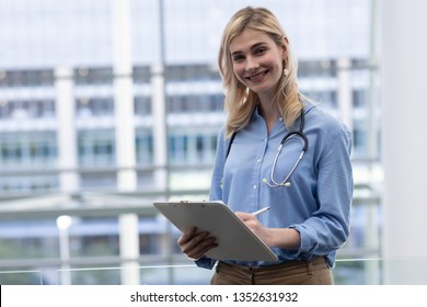 Portrait of beautiful young blonde Caucasian female doctor looking at camera while writing on clipboard standing in clinic