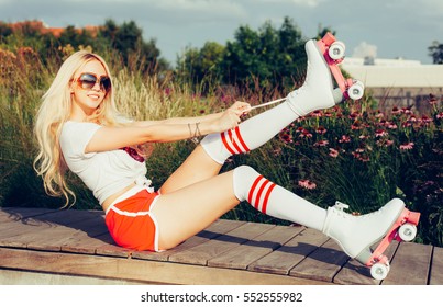 Portrait of a beautiful young blonde girl tighten the laces sitting on a bench in a vintage roller skates, wearing shorts, golfs and a T-shirt. Hot summer day. Outdoor.