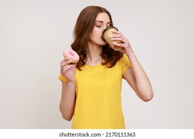 Portrait of beautiful young brown haired teenager girl in casual yellow T-shirt drinking coffee from disposable cup with delicious donut. Indoor studio shot isolated on gray background.