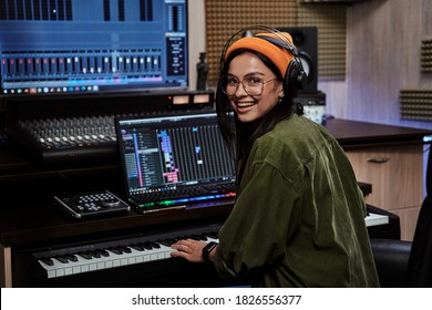 Portrait of beautiful young brunette, female artist smiling at camera while playing keyboard synthesizer, sitting in recording studio - Powered by Shutterstock