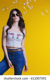 Portrait of beautiful young brunette with glasses in white top and jeans standing on a yellow background in the Studio .Bubbles - Shutterstock ID 611007392