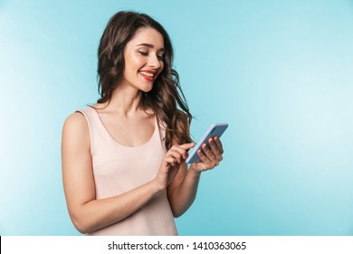 Portrait of a beautiful young brunette woman standing isolated over blue background, using mobile phone