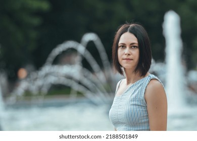 Portrait Of A Beautiful Young Brunette Woman With A Short Bob Haircut On A Warm Day For A Walk Against The Backdrop Of A Fountain