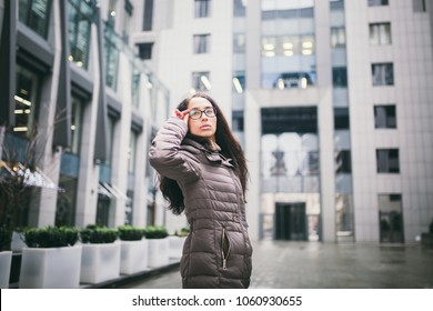 Portrait Beautiful Young Business Woman Brunette In Jacket And Sweater Stands On Background Office Building, Business Center In Glasses For View. Hand Holding Onto Shackle Of Glasses And Looking Away.