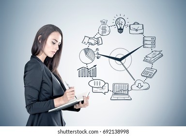 Portrait of a beautiful young businesswoman wearing a black suit and holding a planner and a pen taking notes. Gray wall background with a black time management sketch on it.