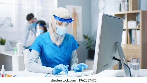 Portrait Of Beautiful Young Busy Arab Female Healthcare Worker Nurse In Medical Mask And Face Shield Sitting At Desk In Cabinet Typing On Computer Working In Hospital. Woman Doctor, Covid Pandemic