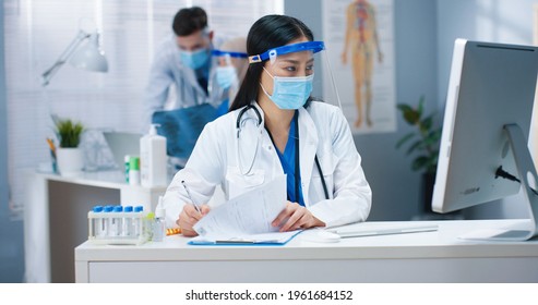 Portrait Of Beautiful Young Busy Asian Female Healthcare Worker In Medical Mask And Face Shield Sitting At Desk In Cabinet Typing On Computer And Writing In Documents. Hospital, Covid Pandemic