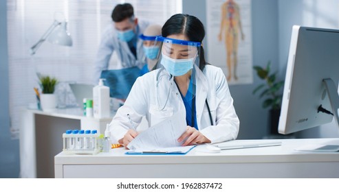 Portrait Of Beautiful Young Busy Asian Female Healthcare Worker In Medical Mask And Face Shield Sitting At Desk In Cabinet Typing On Computer And Writing In Documents. Hospital, Covid Pandemic