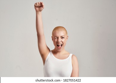 Portrait of a beautiful young caucasian woman with shaved head wearing white shirt, winking at camera and raising clenched fist, standing isolated over grey background. Front view. Horizontal shot