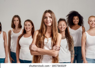 Portrait Of Beautiful Young Caucasian Woman With Long Hair In White Shirt Smiling At Camera. Group Of Diverse Women Posing, Standing Isolated Over Grey Background. Diversity Concept. Selective Focus