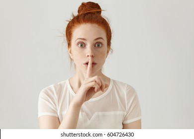 Portrait of beautiful young Caucasian woman with ginger hair bun holding index finger at lips, asking to keep silence or not tell anyone her secret, raising brows, saying "Shh", "Hush", "Tsss"
