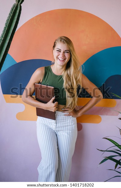 Portrait of a beautiful, young, Caucasian woman in a casual and comfortable outfit. She is holding her laptop computer and is smiling as she basks in the sunlight and poses for her head shot.