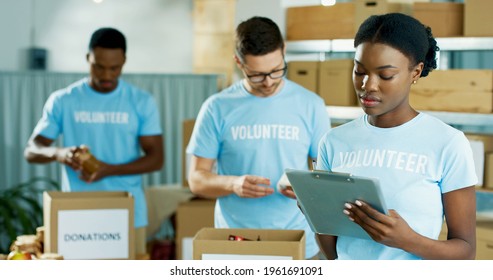 Portrait Of Beautiful Young Concentrated African American Woman Volunteer Standing In Charity Organization Warehouse Writing And Checking Donations. Male Workers Sorting Donated Stuff In Boxes Behind