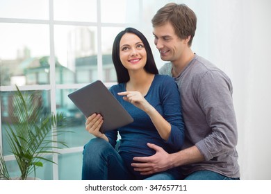 Portrait of beautiful young couple. Man and woman sitting near big window with tablet computer at home. Pregnant woman feeling comfortable and smiling