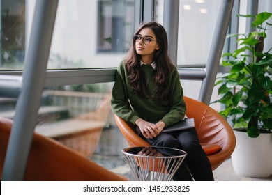 Portrait Of A Beautiful, Young, Elegant And Attractive Indian Asian Woman Student In A Preppy Green Sweater And Spectacles. She Is Relaxing In A Glass Building During The Day. 