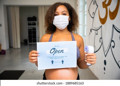 Portrait Of Beautiful Young Female Teacher And Owner In Gym With Face Mask Thermometer In Hand And Sign That Says We Are Open Please Put On Face Mask And Keep Your Distance For Protection Coronavirus