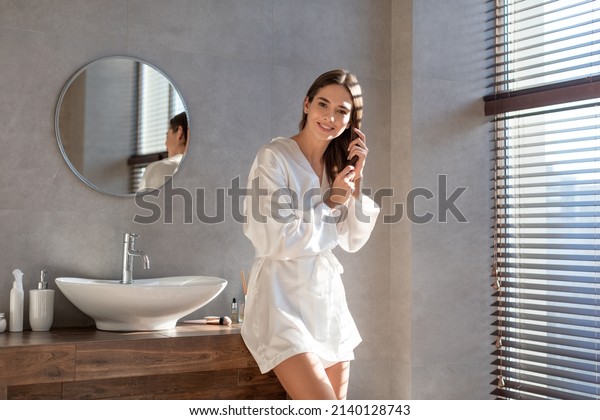 Portrait Of Beautiful Young Female Wearing White\
Silk Robe Posing In Luxury Bathroom Interior, Attractive Millennial\
Woman Making Beauty Treatments At Home, Touching Her Hair And\
Smiling At Camera