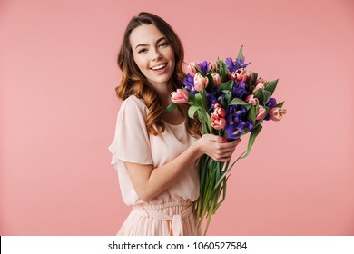 Portrait of a beautiful young girl in dress holding big bouquet of irises and tulips isolated over pink background