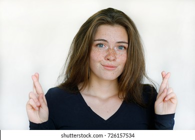 Portrait Of A Beautiful Young Girl With A Freckled Face In A Black Sweatshirt With Curvy Hair Crossed Fingers In Hands Awaiting The Results Of A Sweepstakes Against A Light Wall Background