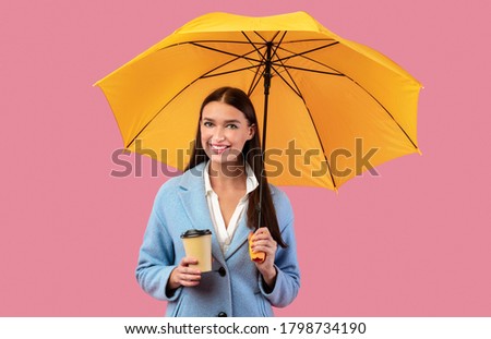 Portrait of beautiful young girl holding yellow umbrella and cup of coffee, isolated over pastel pink wall