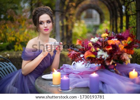 Portrait of a beautiful young girl in a lilac dress drinking tea in the autumn garden at a table with a lilac tablecloth, a bright red bouquet and candles. Romantic evening photo.