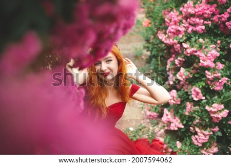 portrait of a beautiful young girl with red hair and a red dress in a garden of roses 