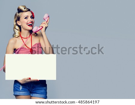 Portrait of beautiful young happy smiling woman with phone and blank signboard, dressed in pin-up style. Caucasian blond model posing in retro fashion and vintage concept studio shoot.