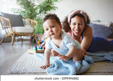 Portrait Of Beautiful Young Hispanic Woman Wearing Headband Babysitting Infant Boy, Watching Him Getting Around By Himself With Confident Smile. Toddler Crawling Along Floor In Bedroom With Mom