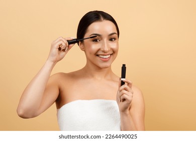 Portrait Of Beautiful Young Indian Female Applying Mascara On Eyelashes, Attractive Hindu Woman Making Daily Makeup And Smiling At Camera, Standing Wrapped In Towel On Beige Background, Copy Space - Powered by Shutterstock