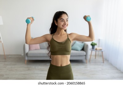 Portrait of beautiful young Indian woman keeping fit, training with dumbbells at home. Sporty Eastern lady performing strength exercises in living room. Staying healthy during covid-19 lockdown