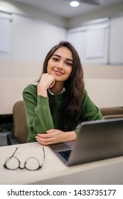 Portrait Of A Beautiful, Young And Intelligent-looking Indian Asian Woman Student Wearing A White Shirt And Green Tracker Smiling As She Works On Her Laptop In A University Classroom. 