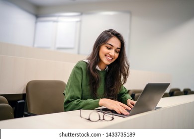 Portrait of a beautiful, young and intelligent-looking Indian Asian woman student wearing a white shirt and green tracker smiling as she works on her laptop in a university classroom. 
