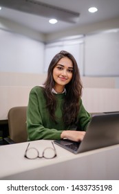 Portrait Of A Beautiful, Young And Intelligent-looking Indian Asian Woman Student Wearing A White Shirt And Green Tracker Smiling As She Works On Her Laptop In A University Classroom. 